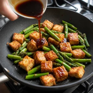 Tofu crocante devolvido à frigideira com aspargos enquanto o molho de gengibre e soja é despejado por cima.
