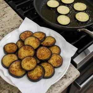 Fried eggplant slices draining on paper towels beside a skillet
