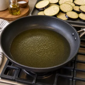 Olive oil heating in a skillet with eggplant slices nearby