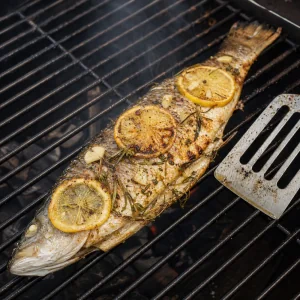 Ganzer gegrillter Fisch auf dem Rost gewendet mit Zitronenscheiben und zerdruecktem Knoblauch darauf