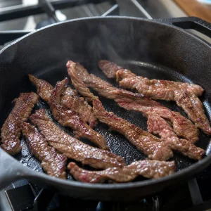 Thin strips of flank steak searing in a hot cast iron skillet
