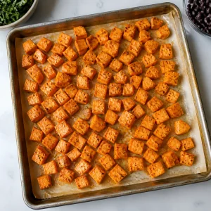 Cubetti di patata dolce conditi su una teglia rivestita di carta forno.