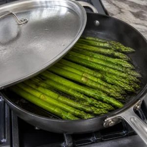 Asparagus steaming in olive oil under a partly covered skillet lid