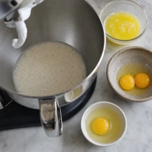 Foamy yeast mixture in a mixer bowl with butter and eggs nearby