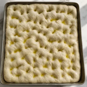 Deep dimples pressed across puffy focaccia dough with olive oil