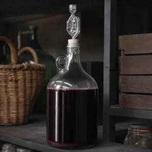 Glass carboy of young red wine sealed with an airlock on a dark shelf