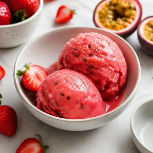 Two scoops of strawberry passion fruit sorbet in a bowl with fresh strawberries and passion fruit.