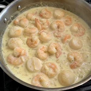 Shrimp and scallops gently simmering in pale cream sauce in a saucepan.