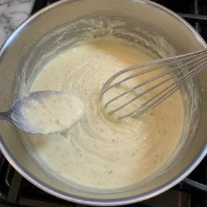 Smooth milk and white wine cream sauce thickening in a saucepan with a whisk and spoon.
